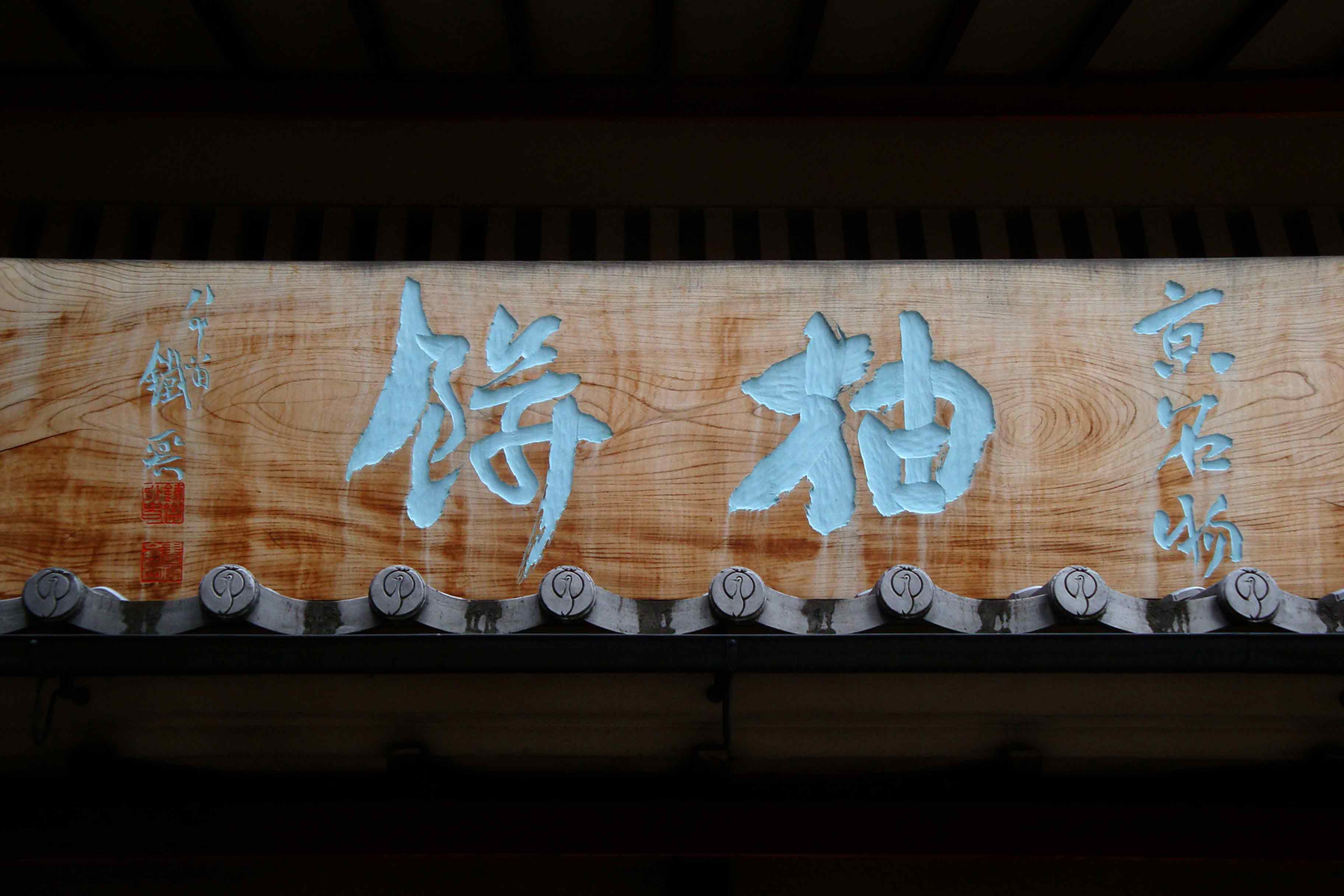 鶴屋吉信本店入口上を飾るのは、樹齢数百年の欅（けやき）の一枚板に緑青胡粉押しの大看板。 文人画家・富岡鉄斎画伯の揮毫です